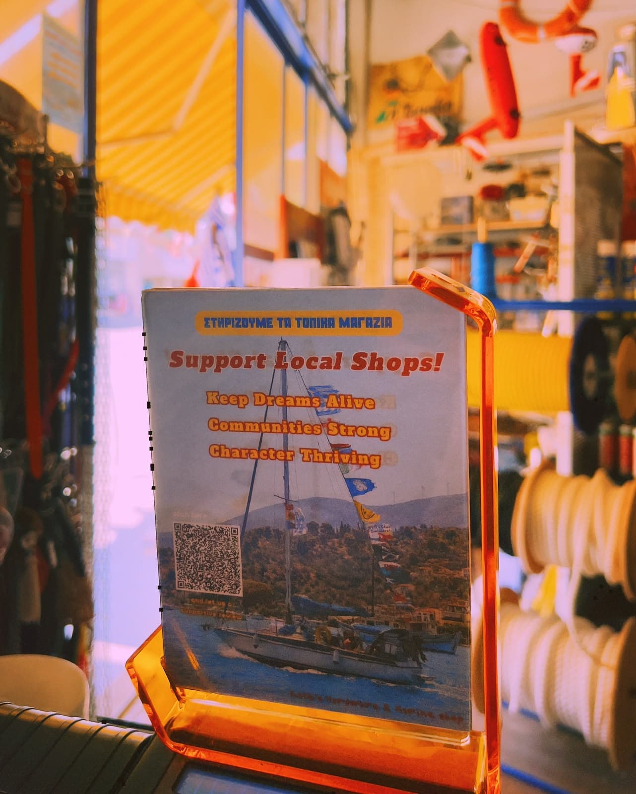 A card on the counter reading “Support Local Shops!”, with a sailing boat and a QR code, rope reels behind.