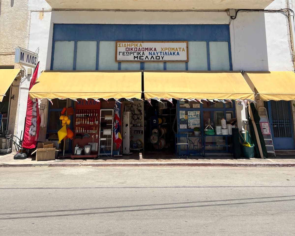 The whole building under a blue sky: white walls, two balconies, the yellow awnings and goods set out on the pavement.