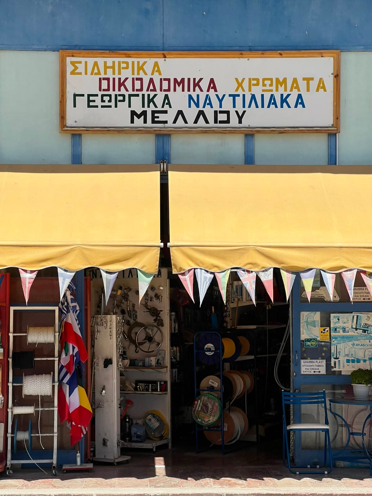 The hand-painted shop sign: ΣΙΔΗΡΙΚΑ, ΟΙΚΟΔΟΜΙΚΑ, ΧΡΩΜΑΤΑ, ΓΕΩΡΓΙΚΑ, ΝΑΥΤΙΛΙΑΚΑ, ΜΕΛΛΟΥ, above the yellow awning, bunting and rope reels at the door.