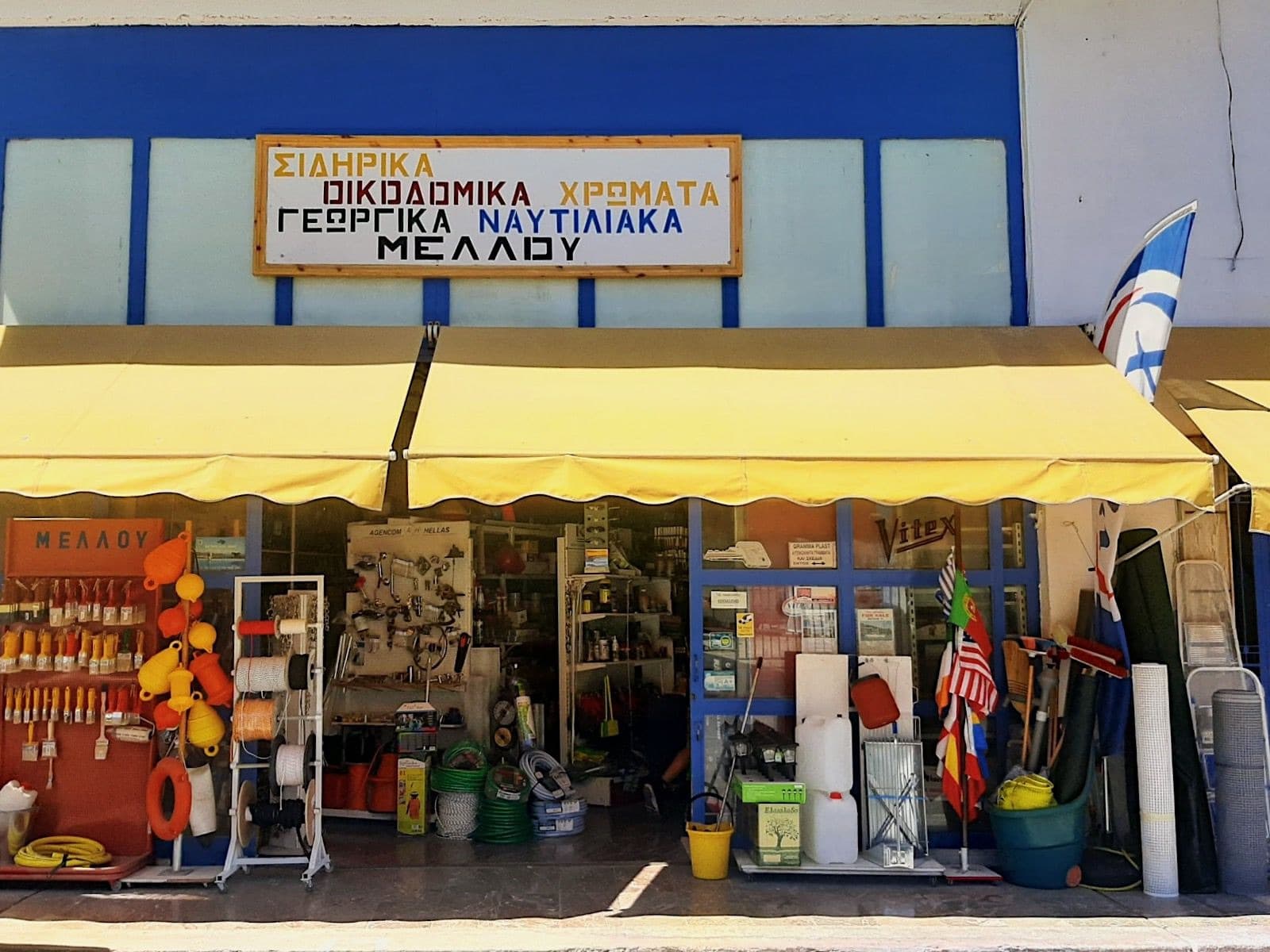 The shopfront on 25 Μαρτίου in Galatas: a yellow awning under a blue frame, the ΜΕΛΛΟΥ sign reading ΣΙΔΗΡΙΚΑ, ΟΙΚΟΔΟΜΙΚΑ, ΧΡΩΜΑΤΑ, ΓΕΩΡΓΙΚΑ, ΝΑΥΤΙΛΙΑΚΑ, and brushes, fenders, rope and hose set out at the front.